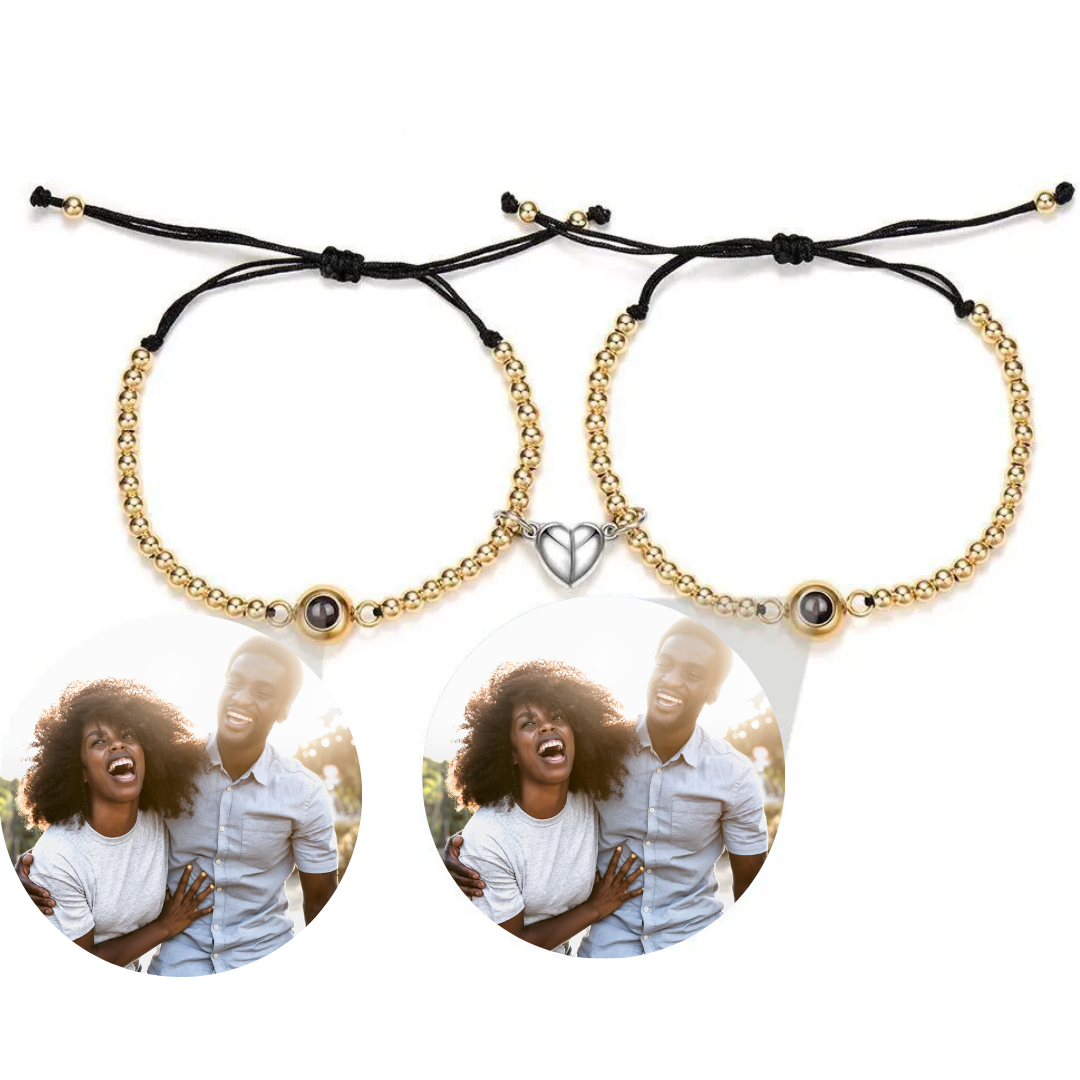 Zwei stilvolle Armbänder mit goldenen Perlen und schwarzer Kordel, die jeweils eine personalisierbare Fotolinse enthalten. Beide Armbänder sind mit einem herzförmigen Anhänger verziert, der in der Mitte geteilt ist und sich magnetisch zusammenhält. Die Fotolinsen zeigen das Bild eines fröhlichen Paares, das gemeinsam lacht. Der schlichte, weiße Hintergrund lässt die Armbänder und die lebhaften Bilder besonders gut zur Geltung kommen.
