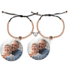 Zwei elegante Armbänder mit roségoldenen Perlen und schwarzer Kordel, die jeweils eine personalisierbare Fotolinse enthalten. Beide Armbänder sind mit einem herzförmigen Anhänger verziert, der in der Mitte geteilt ist und sich magnetisch zusammenhält. Die Fotolinsen zeigen das Bild eines glücklichen älteren Paares, das sich herzlich umarmt. Der schlichte, weiße Hintergrund lässt die Armbänder und die liebevollen Bilder besonders gut zur Geltung kommen.