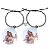Zwei schwarze Armbänder mit Perlen und schwarzer Kordel, die jeweils eine personalisierbare Fotolinse enthalten. Beide Armbänder sind mit einem herzförmigen Anhänger verziert, der in der Mitte geteilt ist und sich magnetisch zusammenhält. Die Fotolinsen zeigen das Bild eines glücklichen jungen Paares in einer liebevollen Umarmung. Der Hintergrund  ist neutral.