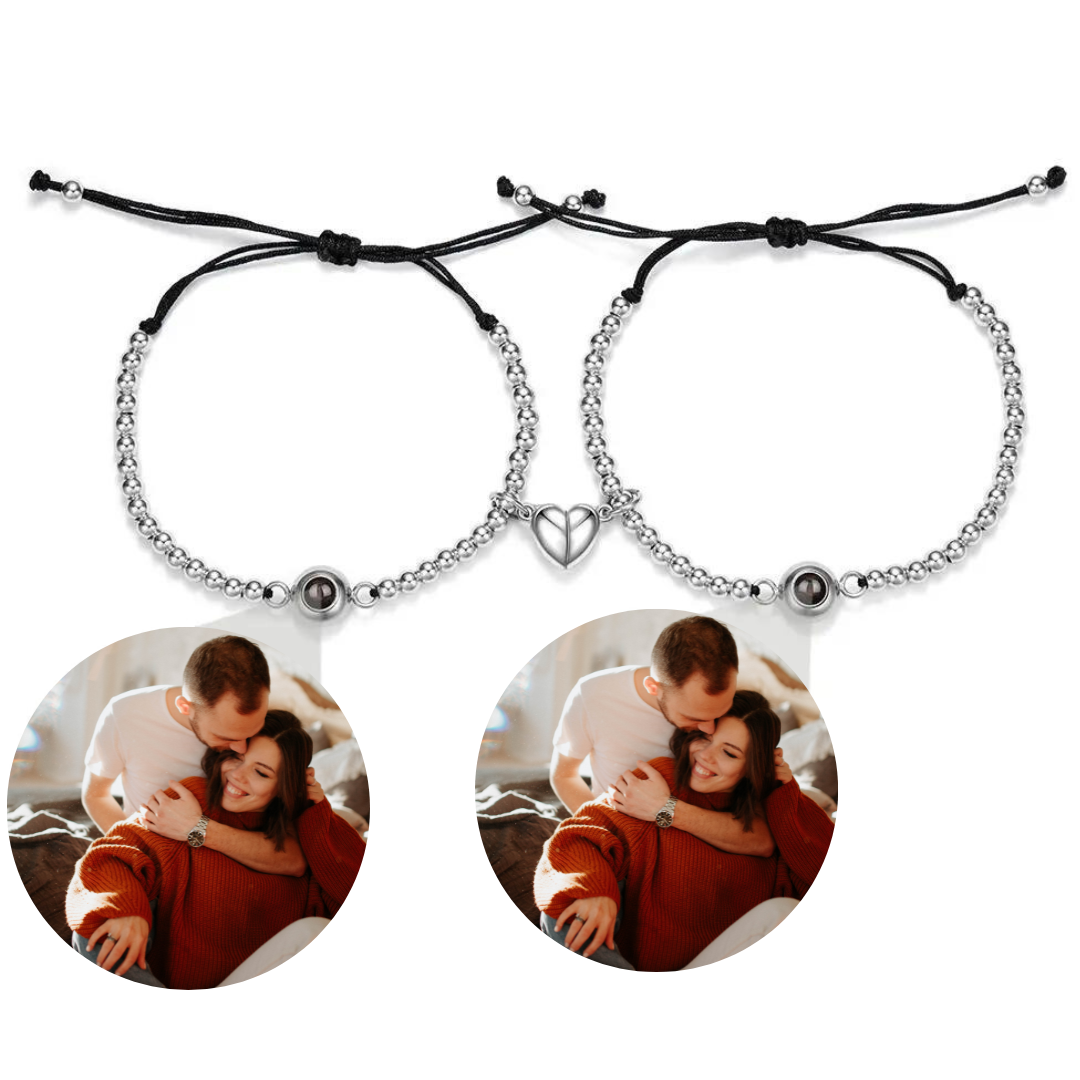Zwei silberne Armbänder mit Perlen und schwarzer Kordel, die jeweils eine personalisierbare Fotolinse enthalten. Beide Armbänder sind mit einem herzförmigen Anhänger verziert, der in der Mitte geteilt ist und sich magnetisch zusammenhält. Die Fotolinsen zeigen das Bild eines Paares, das sich in einer liebevollen Umarmung befindet. Der Hintergrund ist neutral.