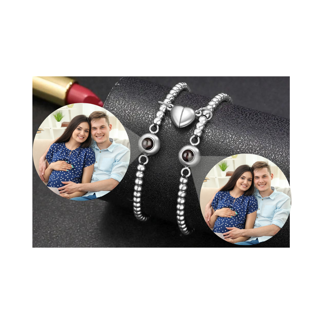 Zwei elegante Armbänder mit silbernen Perlen und personalisierbaren Fotolinsen, die ein glückliches Paar zeigen. Jedes Armband ist mit einem herzförmigen Magnetverschluss verziert, der die beiden Armbänder zusammenhält. Das Bild zeigt die Armbänder auf einem schwarzen, texturierten Hintergrund, während die Fotolinsen das Bild eines werdenden Elternpaares in einer gemütlichen Umgebung zeigt. Diese Armbänder sind eine besondere Erinnerung an die gemeinsamen Momente und die Liebe zwischen den Partnern.