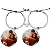 Zwei silberne Armbänder mit Perlen und schwarzer Kordel, die jeweils eine personalisierbare Fotolinse enthalten. Beide Armbänder sind mit einem herzförmigen Anhänger verziert, der in der Mitte geteilt ist und sich magnetisch zusammenhält. Die Fotolinsen zeigen das Bild eines Paares, das sich in einer liebevollen Umarmung befindet. Der Hintergrund ist neutral.