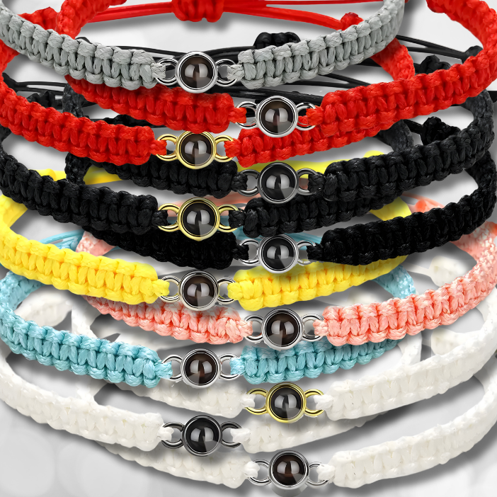 Eine Auswahl geflochtener Armbänder in verschiedenen Farben, darunter Grau, Rot, Schwarz, Gelb, Türkis, Weiß und Rosa, alle mit goldenen und silbernen Charms in der Mitte, der eine personalisierbare Fotolinse enthält. Die Armbänder sind übereinander gestapelt und auf einem hellen Hintergrund angeordnet.