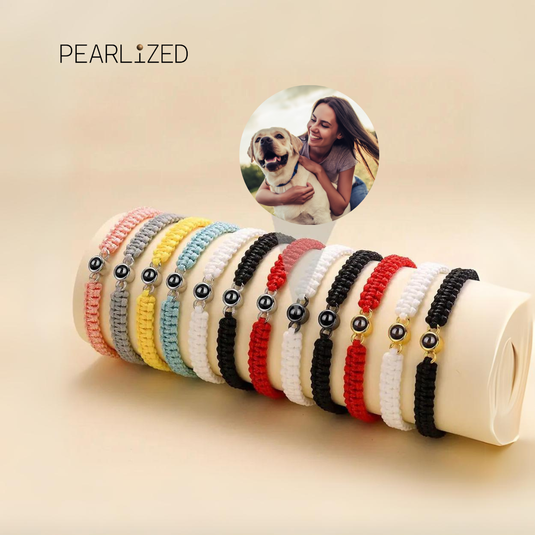 Auswahl an Makramee-Armbändern in verschiedenen Farben (Rosa, Grau, Gelb, Hellblau, Schwarz, Rot, Weiß) mit einer Fotoperle / Fotolinse in der Mitte, präsentiert auf einer Rolle. Beispielprojektion zeigt eine Frau, die einen Hund umarmt. Pearlized-Logo im Hintergrund.
