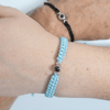 Zwei Handgelenke mit geflochtenen Armbändern, eines in hellblau mit silbernem Charm und das andere in schwarz mit silbernem Charm. Beide Armbänder haben eine personalisierbare Fotolinse in der Mitte. Heller Hintergrund.