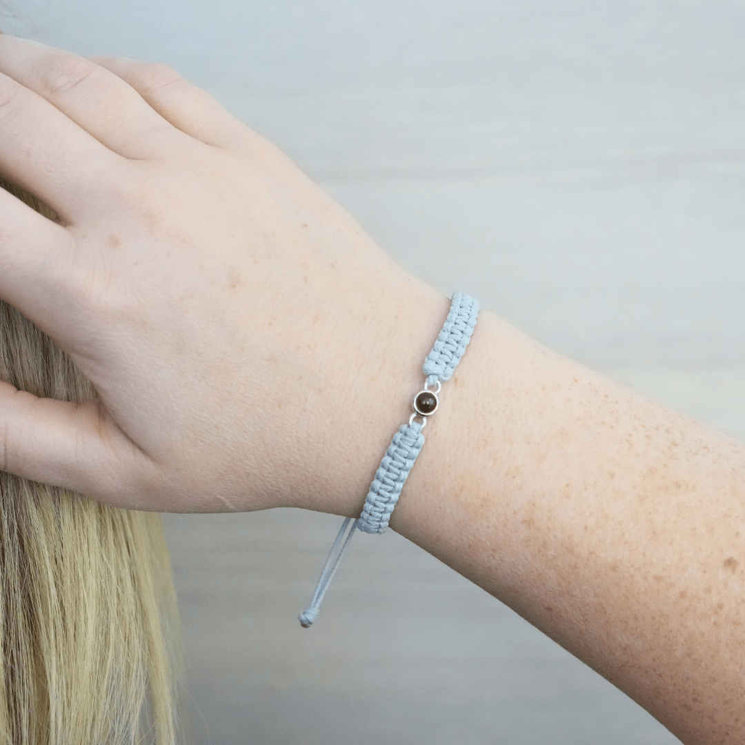 Handgelenk mit einem grauen geflochtenen Armband und silbernem Charm in der Mitte, der eine personalisierbare Fotolinse enthält. Der Hintergrund ist hell und eine Hand greift ins Haar.