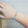 Handgelenk mit einem grauen geflochtenen Armband und silbernem Charm in der Mitte, der eine personalisierbare Fotolinse enthält. Der Hintergrund ist hell und eine Hand greift ins Haar.