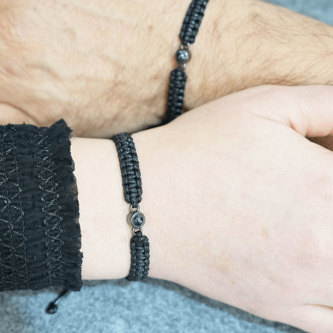 Zwei Handgelenke, jeweils mit einem schwarzen Makramee-Armband mit einer Fotoperle / Fotolinse in der Mitte. Heller Hintergrund.