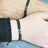 Zwei Handgelenke, jeweils mit einem schwarzen Makramee-Armband mit einer Fotoperle / Fotolinse in der Mitte. Heller Hintergrund.