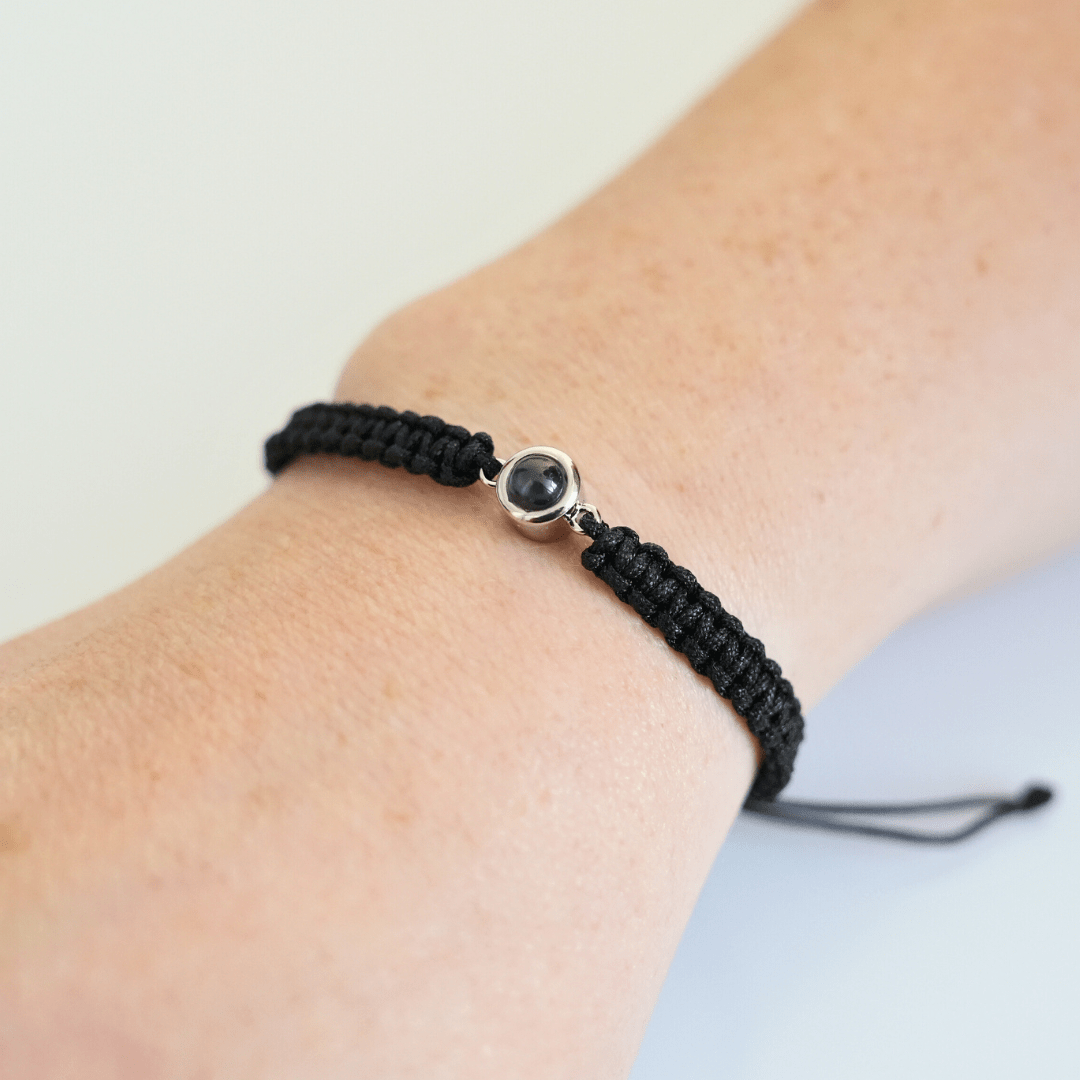 Handgelenk mit einem schwarz geflochtenen Armband und silbernem Charm in der Mitte, der eine personalisierbare Fotolinse enthält. Der Hintergrund ist hell.