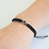 Handgelenk mit einem schwarz geflochtenen Armband und silbernem Charm in der Mitte, der eine personalisierbare Fotolinse enthält. Der Hintergrund ist hell.