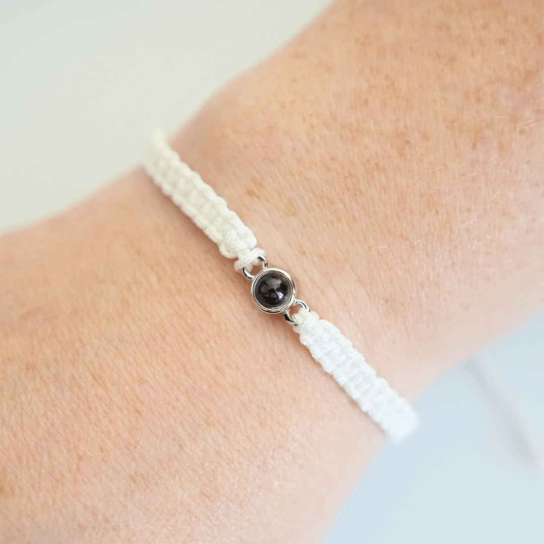 Handgelenk mit einem weiß geflochtenen Armband und silbernem Charm in der Mitte, der eine personalisierbare Fotolinse enthält. Der Hintergrund ist hell.
