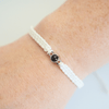 Handgelenk mit einem weiß geflochtenen Armband und silbernem Charm in der Mitte, der eine personalisierbare Fotolinse enthält. Der Hintergrund ist hell.
