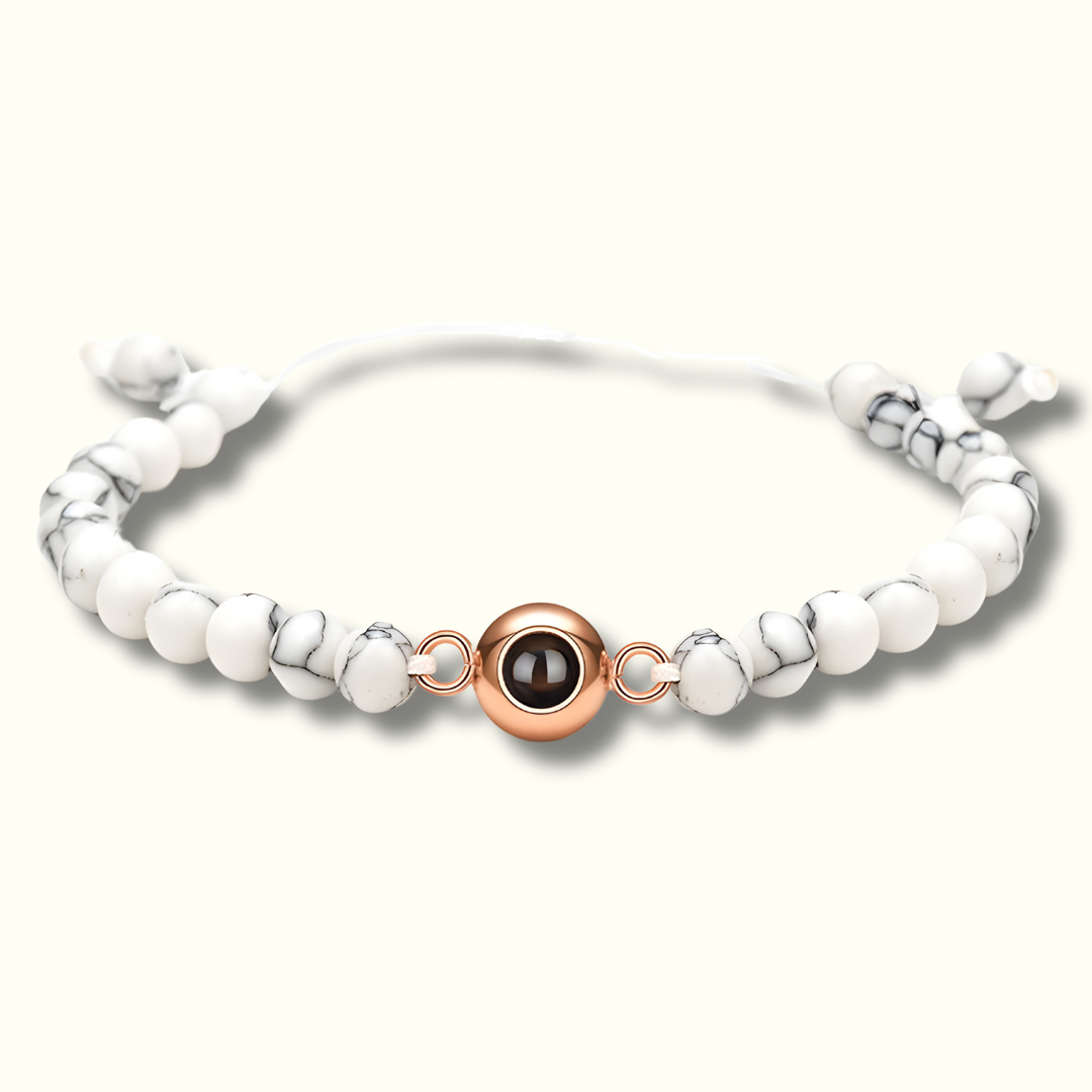 Ein Armband mit weißen Perlen mit einem leichten Marmoreffekt in dunkel bis schwarz. In der Mitte befindet sich ein rosegoldener Charm mit einer Fotoperle / Fotolinse. Der Hintergrund ist hell.