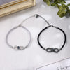 Zwei Infinity-Armbänder auf einer texturierten Unterlage, eines in Silber mit grauem Band und das andere in Schwarz, beide mit einer zentralen Fotolinse für personalisierte Bilder.
